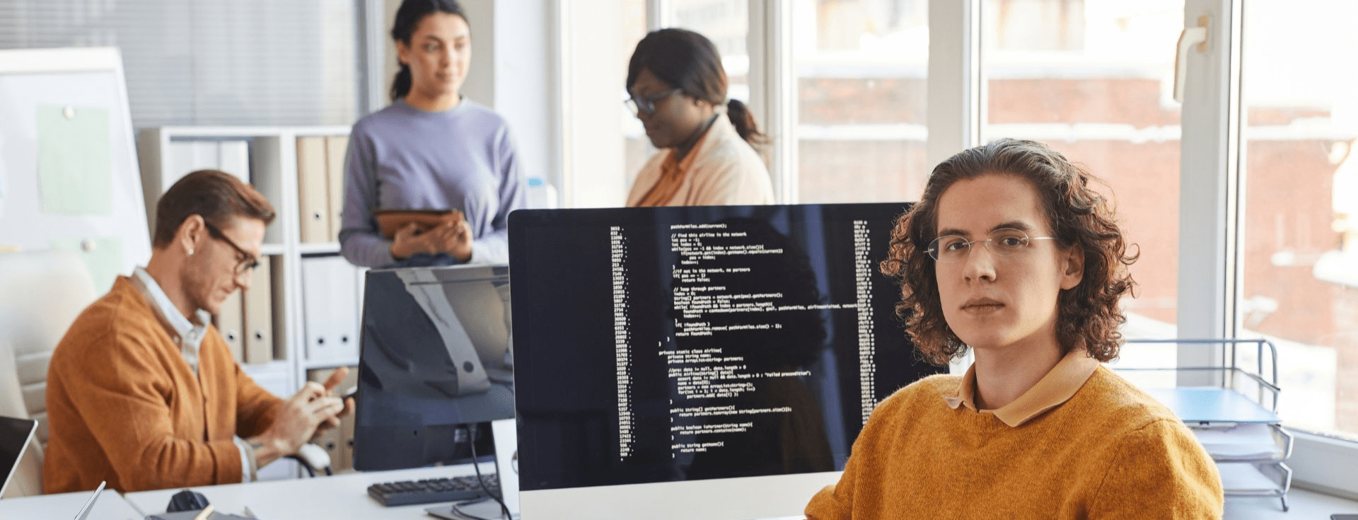 A group of people is gathered in an office setting. They are engaged in various activities, utilizing computers and laptops. The environment is indoors, likely within an office building, and includes elements like walls and windows. The individuals are dressed in professional clothing, suggesting a work-related context focused on learning or collaboration.